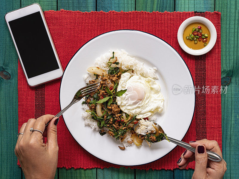著名的泰国辣菜“Pak Krapao Moo”，英文中是用圣罗勒炸猪肉，在一个古老的绿松石色的木桌上，用蒸米饭和煎蛋和“Pik Nam Bla”(辣椒和鱼酱)，一位泰国妇女正在吃。